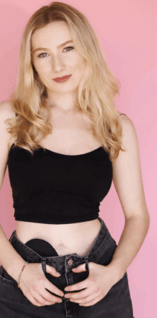 Photo of Keely a white woman with blond hair wearing a black midriff shirt with a black ostomy pouch just visible above the waistline of her black pants. 